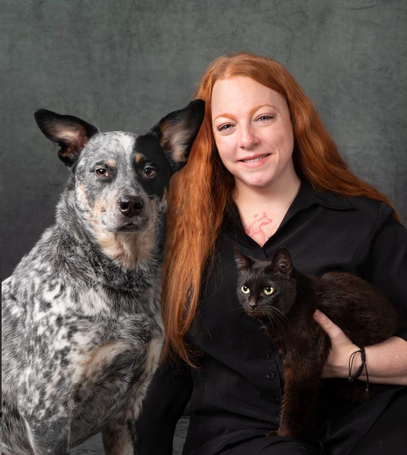 http://Red%20haired%20woman%20wearing%20black%20top%20holding%20a%20black%20cat%20and%20a%20tricolored%20dog%20sitting%20besides%20her.